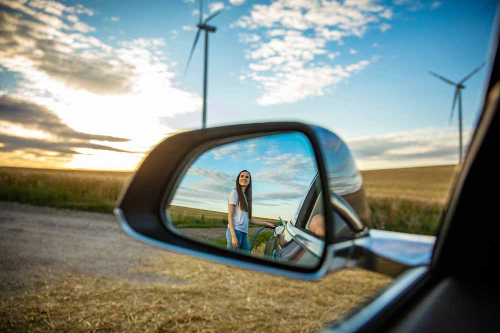 Kvinna laddar elbil medan hon tittar upp på en vindturbin.